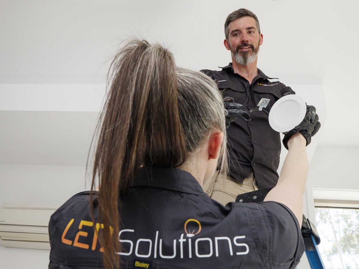Kieran on a ladder fitting an LED downlight while a team member in an LED Solutions shirt hands up the next one