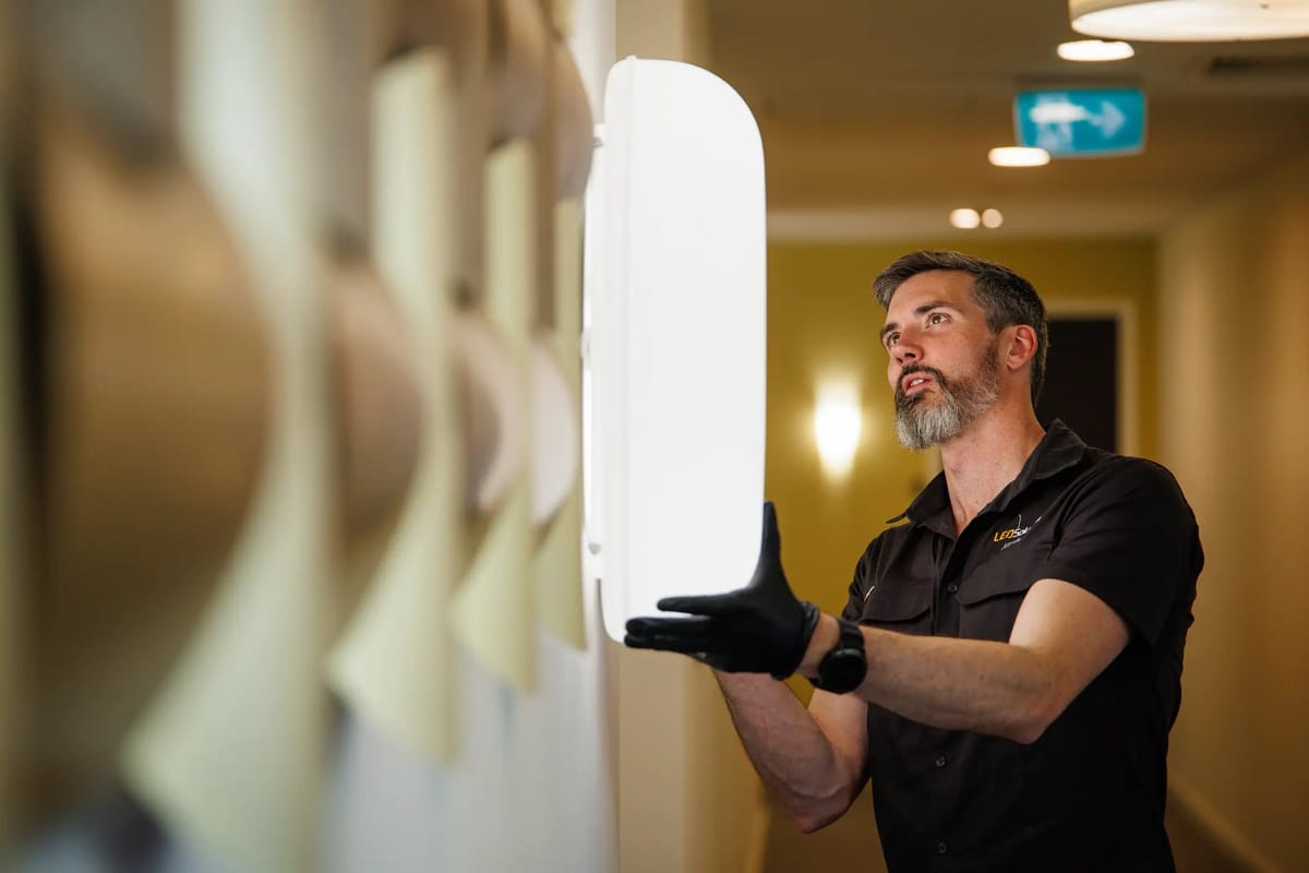 Kieran from LED Solutions fitting a large wall light in a building corridor, exit sign behind
