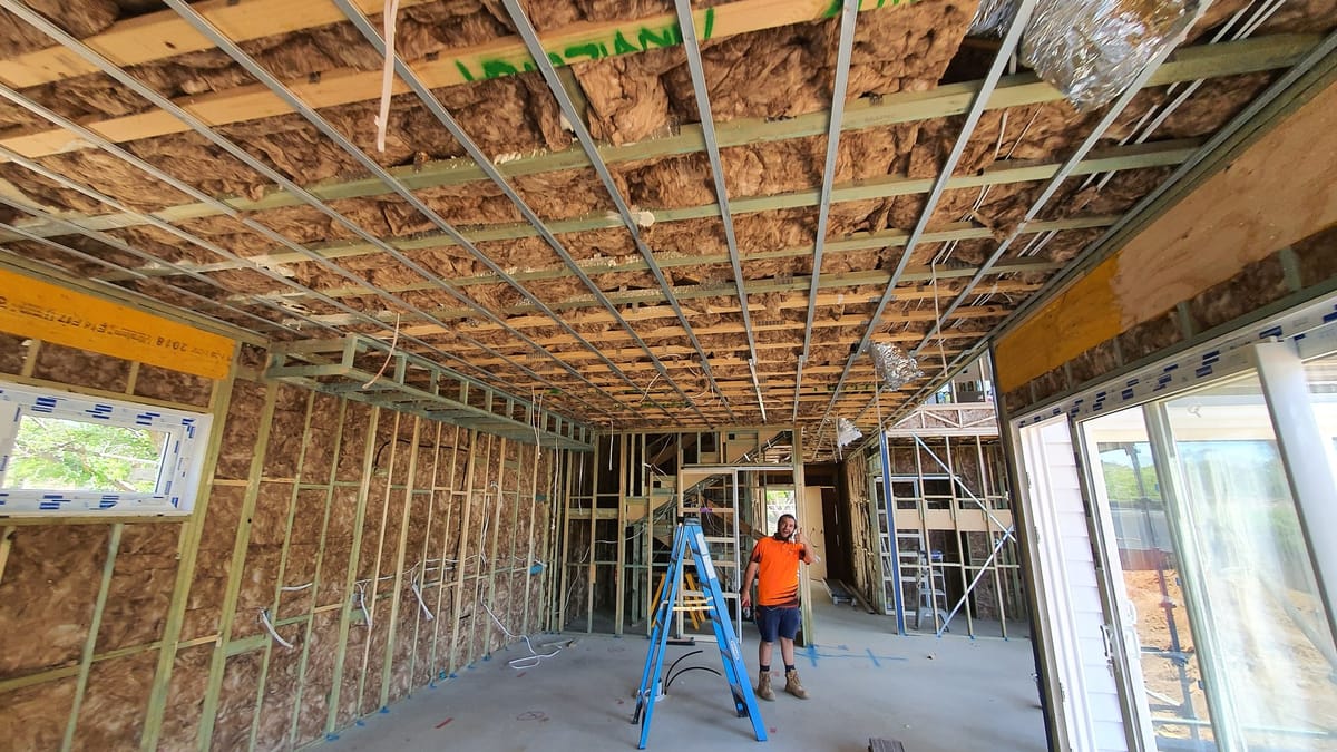 A home at rough-in stage: ceiling battens and insulation with cable drops for the lights, wired timber wall frames, a ladder and a tradesperson on site