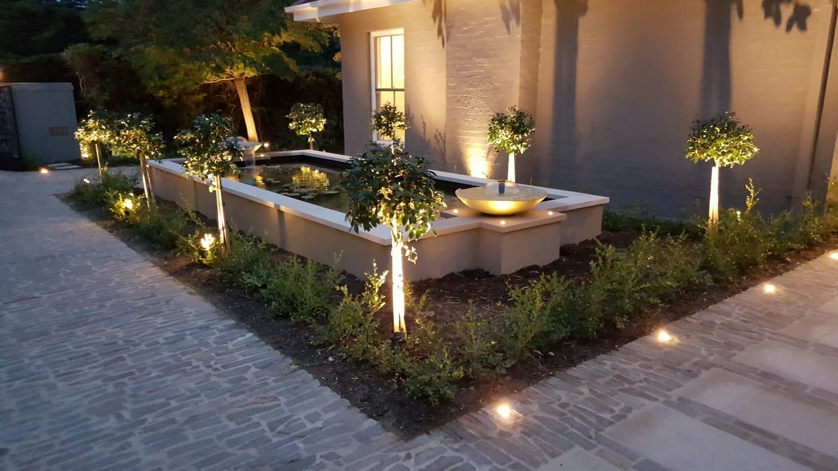 A courtyard garden at dusk: standard trees uplit around a raised pond, a lit water bowl and lights set into the paving