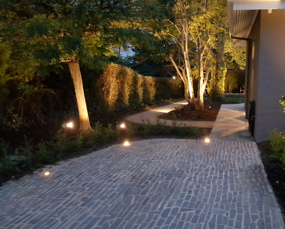 A paved side path at dusk with lights set into the paving and the trees and hedge uplit beside the house
