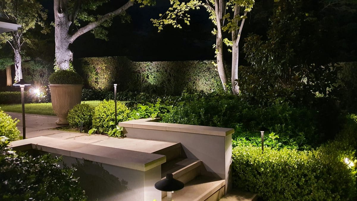 A garden at night with low path lights beside the steps and the trees and hedges gently uplit