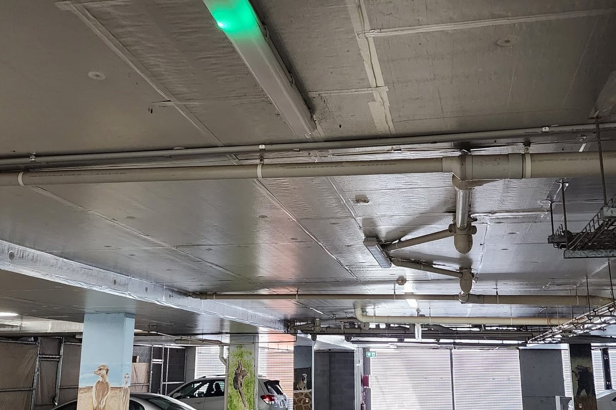 Pipes, a cable tray and a batten light with a green emergency indicator under the ceiling of a Canberra strata car park